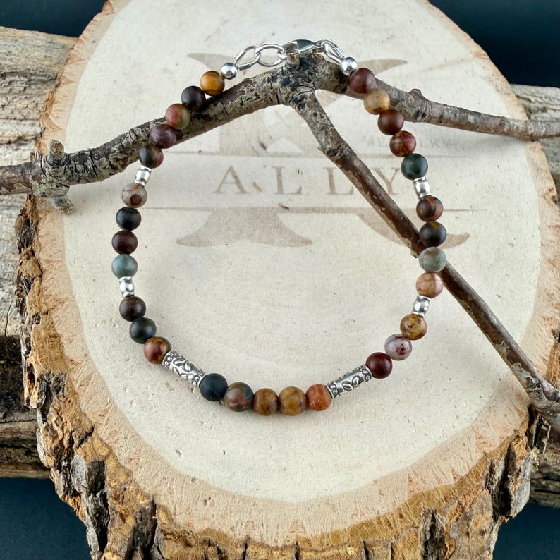 May include: A beaded bracelet featuring round matte earth-toned stones in shades of brown, tan, and muted green, interspersed with small silver-colored metal spacer beads and two ornate silver-colored cylindrical accent beads.