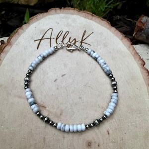May include: A beaded bracelet featuring white and gray stone beads interspersed with silver-colored metal beads. The bracelet is secured with a silver-colored clasp. The word "Allyk" is written in cursive on the wooden surface.