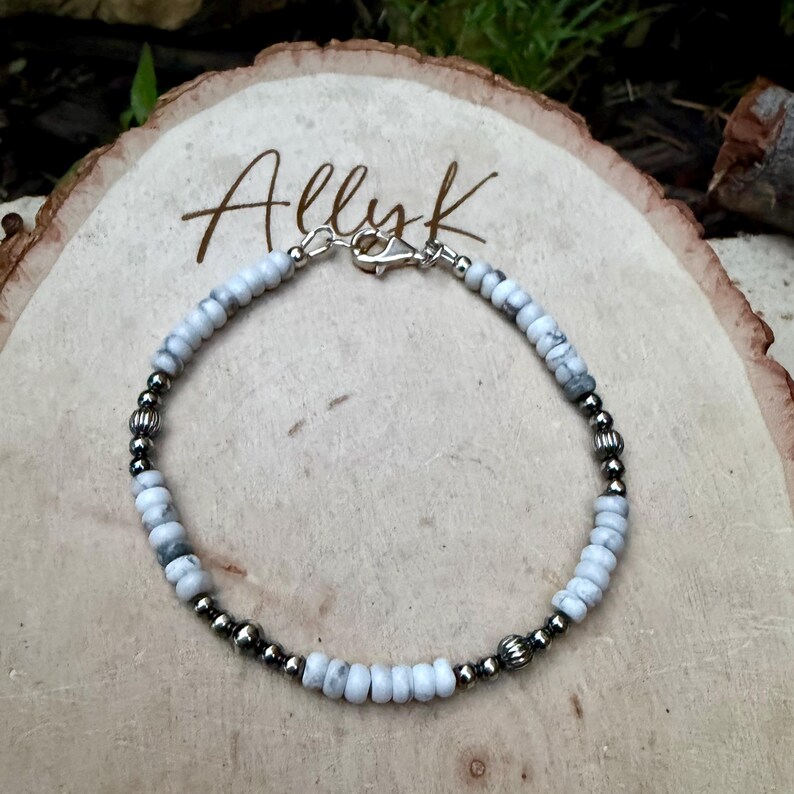 May include: A beaded bracelet featuring white and gray stone beads interspersed with silver-colored metal beads. The bracelet is secured with a silver-colored clasp. The word "Allyk" is written in cursive on the wooden surface.