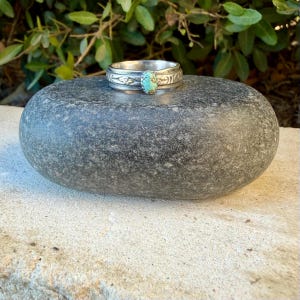 Puede incluir: Un anillo de plata con una piedra turquesa ovalada central con patrones de matriz oscuros. La banda est&aacute; elaborada con un motivo floral decorativo en relieve que rodea toda la circunferencia, aportando un acabado texturizado al metal.