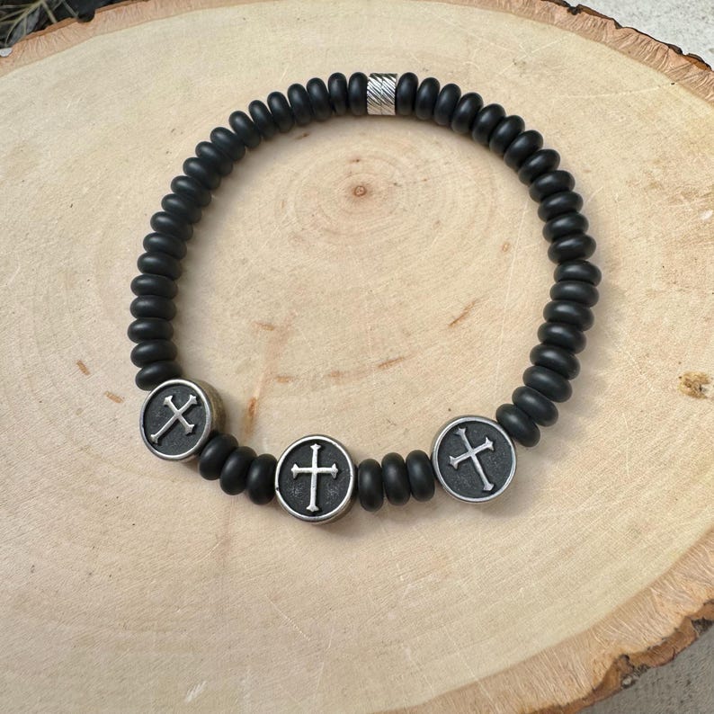 May include: Black beaded bracelet with three silver-colored cross charms. The bracelet features a mix of round and cylindrical beads. A silver-colored cylindrical bead is also present. The bracelet is displayed on a wooden surface.