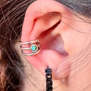 May include: A silver ear cuff with three bands and a turquoise stone in the center. A black chain earring hangs below the ear. The jewelry is worn on the ear.