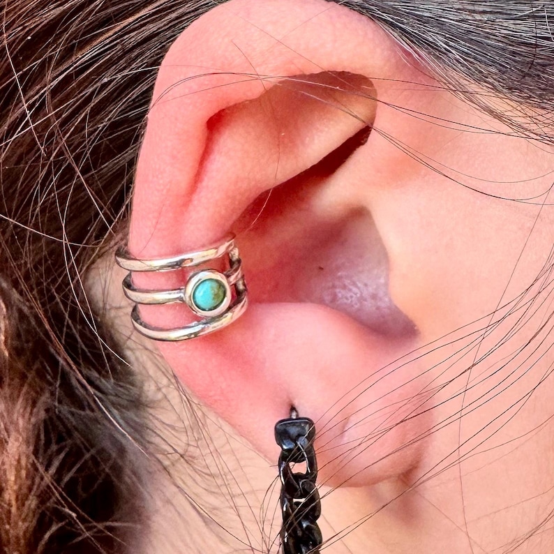 May include: A silver ear cuff with three bands and a turquoise stone in the center. A black chain earring hangs below the ear. The jewelry is worn on the ear.