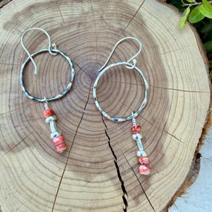 May include: A pair of silver-toned earrings featuring a hammered circular hoop design. Each earring includes a dangling vertical accent composed of two small, mottled orange-red beads separated by a single smooth silver-toned cylindrical spacer.