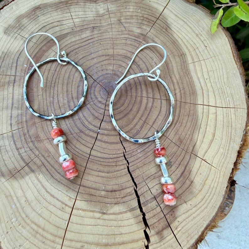 May include: A pair of silver-toned earrings featuring a hammered circular hoop design. Each earring includes a dangling vertical accent composed of two small, mottled orange-red beads separated by a single smooth silver-toned cylindrical spacer.