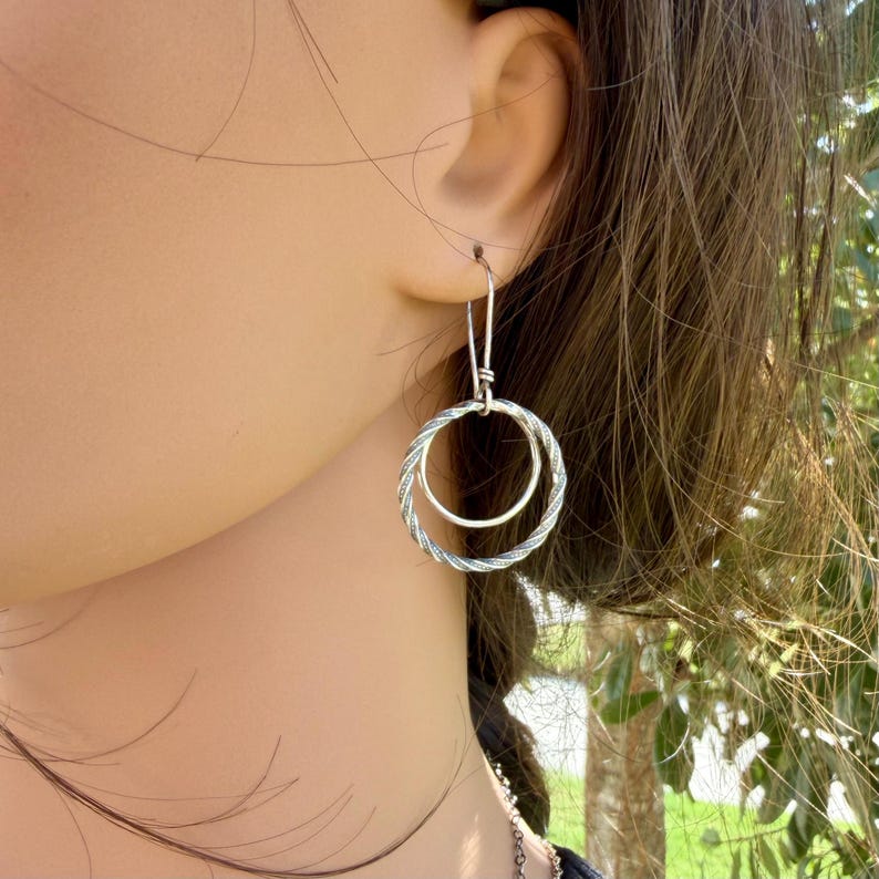 May include: A silver-toned earring featuring two interlocking circular hoops. The larger outer hoop has a twisted, rope-like texture, while the smaller inner hoop is smooth and plain. The earring hangs from a simple, thin silver-toned wire hook.