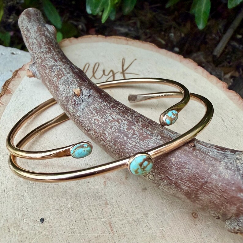 May include: Two gold-colored metal cuff bracelets with turquoise-colored stones. The bracelets are resting on a wooden branch and a wooden surface. The bracelets have a curved design.