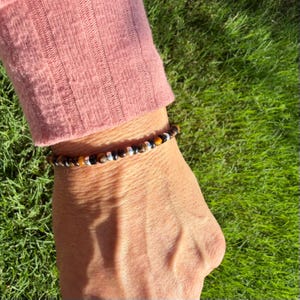 K&ouml;nnte beinhalten: Ein Perlenarmband mit abwechselnd runden Tigerauge-Perlen, glatten schwarzen Perlen und kleinen metallisch silberfarbenen Zwischenelementen. Das Armband wird am Handgelenk getragen, wobei die Perlen in einem sich wiederholenden Muster angeordnet sind.