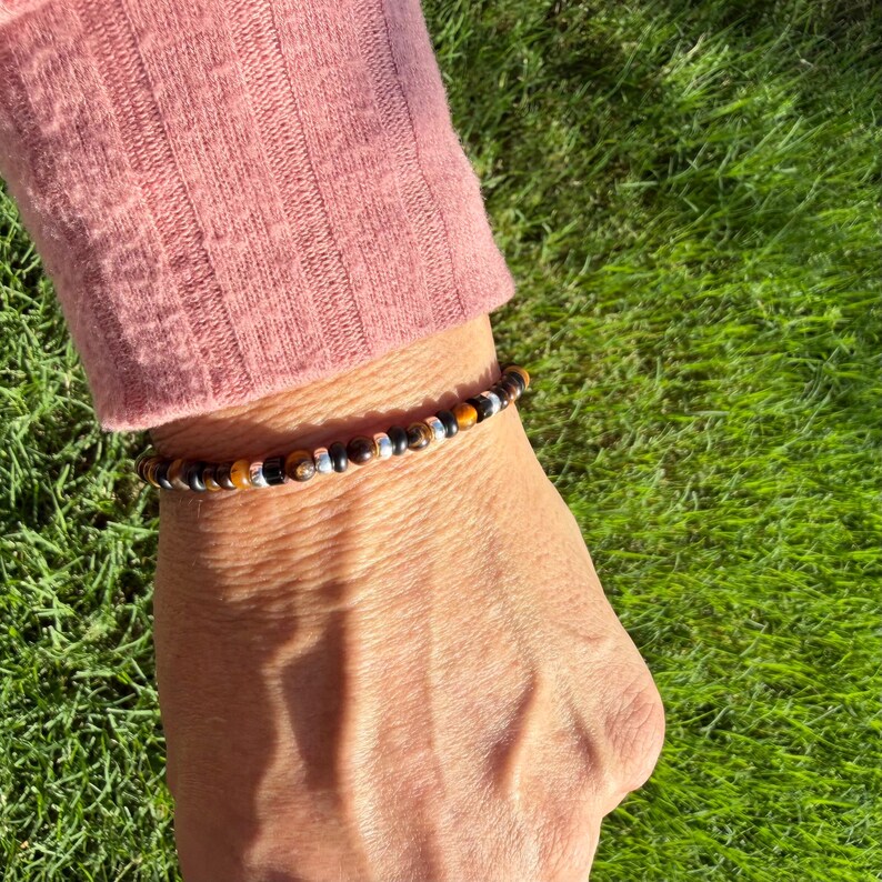 K&ouml;nnte beinhalten: Ein Perlenarmband mit abwechselnd runden Tigerauge-Perlen, glatten schwarzen Perlen und kleinen metallisch silberfarbenen Zwischenelementen. Das Armband wird am Handgelenk getragen, wobei die Perlen in einem sich wiederholenden Muster angeordnet sind.