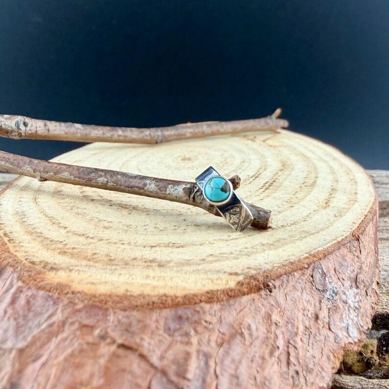 K&ouml;nnte beinhalten: Ein silberner Ring mit einem t&uuml;rkisfarbenen Stein in einem geometrischen Design. Der Ring wird auf einem Holzast pr&auml;sentiert, der seine komplizierten Details und seine Handwerkskunst zeigt. Das Design des Rings weist einen zentralen t&uuml;rkisfarbenen Stein auf.