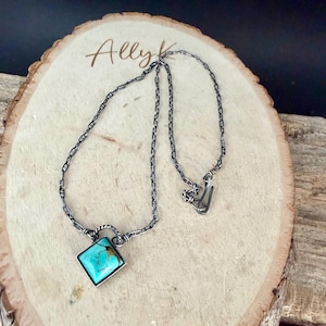 May include: A silver-toned necklace with a square turquoise pendant. The pendant is set in a silver frame and hangs from a chain with a unique link design. The necklace is displayed on a wooden surface.