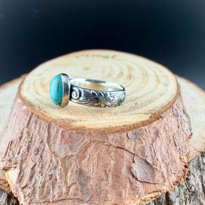 May include: A silver ring with a turquoise stone and floral design. The oval-shaped turquoise stone is set in a silver bezel. The band features an engraved floral pattern. The ring is displayed on a wooden surface.