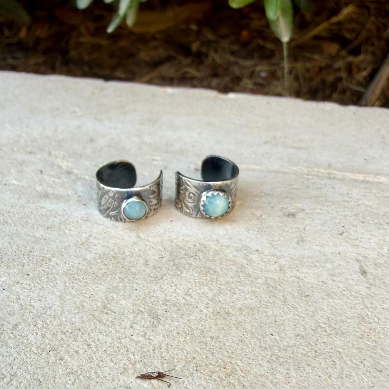 May include: Two open-band rings crafted from oxidized silver, each featuring a round, light blue cabochon stone set in a serrated bezel. The exterior surface of both bands displays an intricate, embossed floral pattern across the metal.