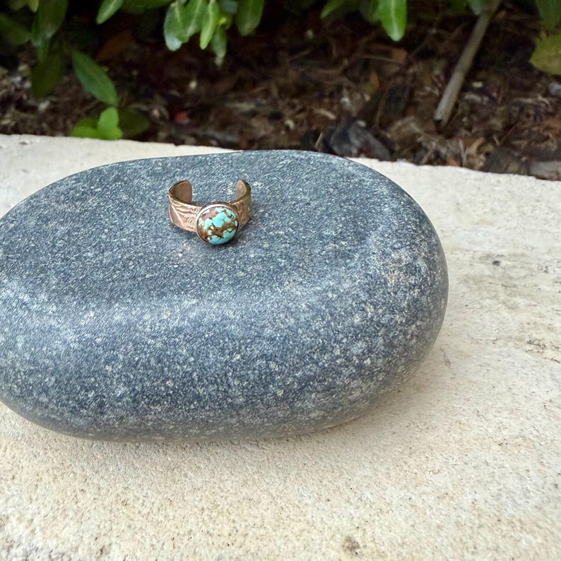 May include: A copper-colored ring with a turquoise stone set on a gray stone. The ring has a textured band with a floral design. The turquoise stone is round and has a mottled appearance.