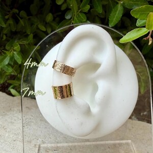 May include: Two gold-colored ear cuffs, 4mm and 7mm wide, are displayed on a white ear model. The cuffs have an engraved floral pattern. The background is blurred, with green foliage visible.