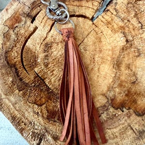 May include: A brown leather tassel keychain with a silver-colored clasp and ring. The tassel is made of long, thin strips of leather. The keychain is shown against a textured, wooden background.