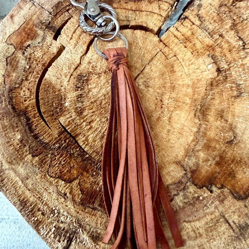 May include: A brown leather tassel keychain with a silver-colored clasp and ring. The tassel is made of long, thin strips of leather. The keychain is shown against a textured, wooden background.