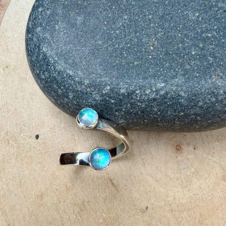 May include: A silver ring featuring two iridescent blue-green gemstones. The ring has an open design, with one stone at the top and another at the bottom. The ring is displayed on a light brown surface. The gemstones are approximately 0.5 cm in diameter.