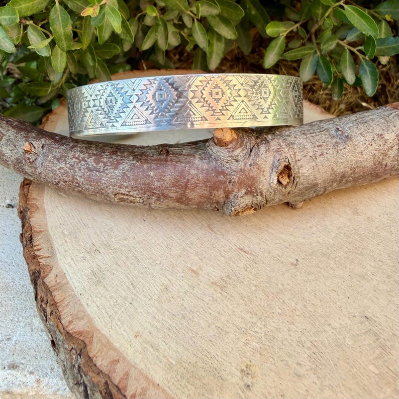 May include: A wide silver-toned metal cuff bracelet featuring a repeating engraved pattern of geometric shapes, including diamonds, triangles, and small dots, creating a textured surface across the entire outer band of the jewelry piece.