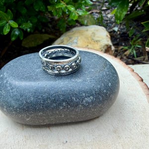 May include: A stack of three silver rings with intricate floral and beaded designs. The rings are displayed on a smooth, gray stone, with a blurred green background of foliage and a light-colored wooden surface.