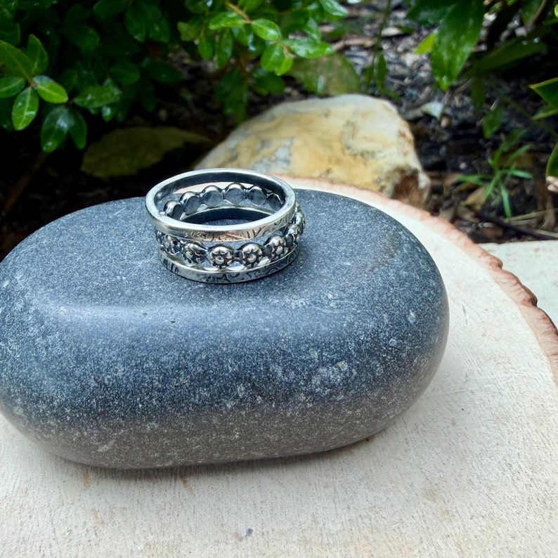 May include: A stack of three silver rings with intricate floral and beaded designs. The rings are displayed on a smooth, gray stone, with a blurred green background of foliage and a light-colored wooden surface.
