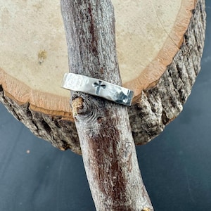 May include: A silver ring with a hammered texture and a four-leaf clover design. The ring is displayed on a wooden branch against a wood slice background. The ring's design is simple and elegant.