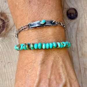 May include: A two-piece bracelet set featuring a silver-toned chain bracelet with a central hammered metal bar and a small turquoise stone, paired with a second bracelet composed of round, vibrant turquoise beads with brown matrix inclusions.