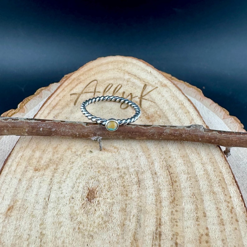 May include: A silver-toned ring with a twisted band and a small, round, light-colored stone. The ring is displayed on a wooden surface with the name "Ally K" lightly etched into the wood.