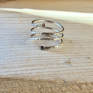 May include: A silver-colored metal ring featuring a spiral design with three distinct coils. The surface of the metal band has a hammered, textured finish that catches the light, creating a series of small indentations along the entire length.