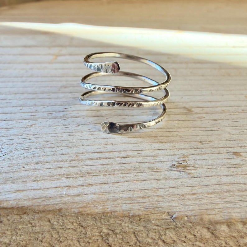 May include: A silver-colored metal ring featuring a spiral design with three distinct coils. The surface of the metal band has a hammered, textured finish that catches the light, creating a series of small indentations along the entire length.