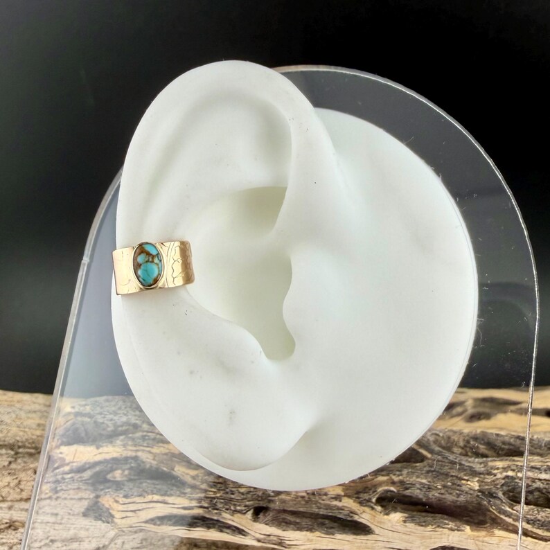 May include: A gold-colored ear cuff with a turquoise stone. The cuff is rectangular with a textured design and is displayed on a white ear model. The turquoise stone is oval-shaped and has a mottled pattern of blue and green.