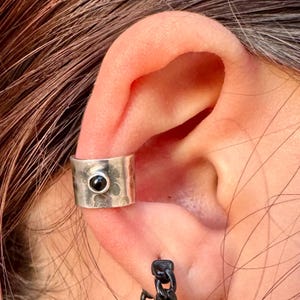 May include: A silver ear cuff with a hammered texture and a small, round black stone set in the center. The cuff is worn on the ear, showcasing a minimalist design. The jewelry is a stylish accessory.