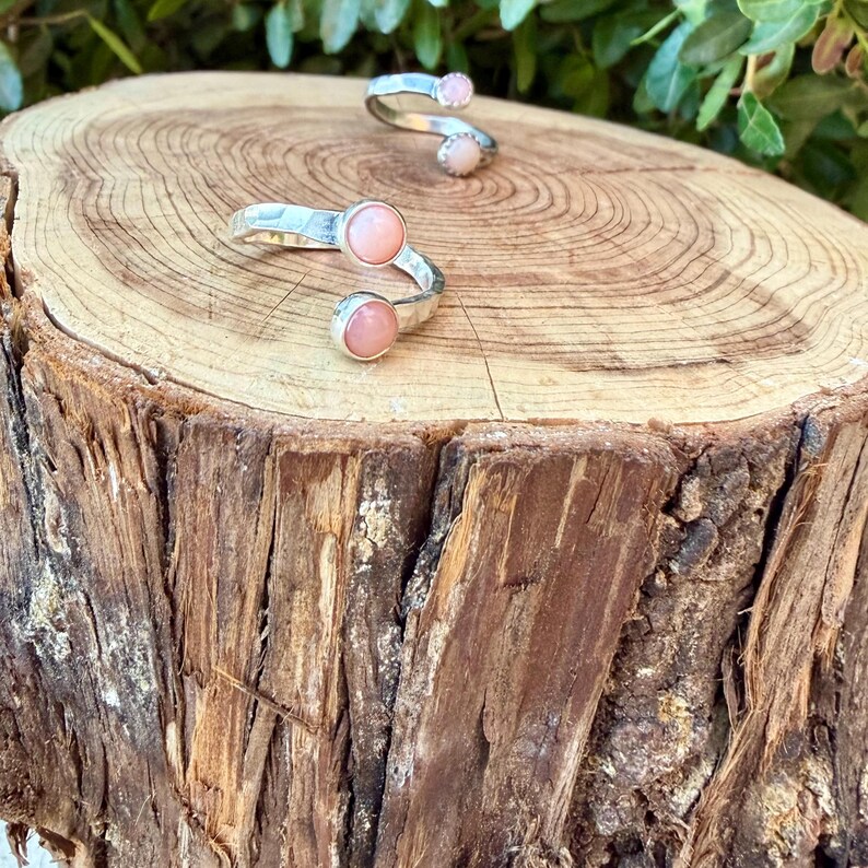 May include: Two open-style rings crafted from polished silver metal, each featuring two round, smooth, pale pink cabochon stones set at the ends of the band. The rings have a bypass design where the ends do not meet, showcasing a clean, metallic finish.