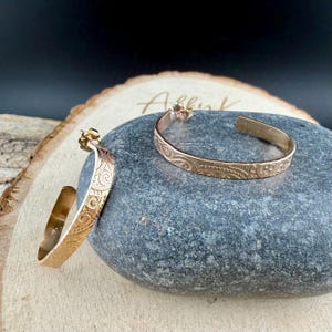 May include: A pair of gold-colored hoop earrings with an engraved floral design. The earrings are displayed on a gray stone, with a wooden background. The earrings have a post and are open at the bottom.