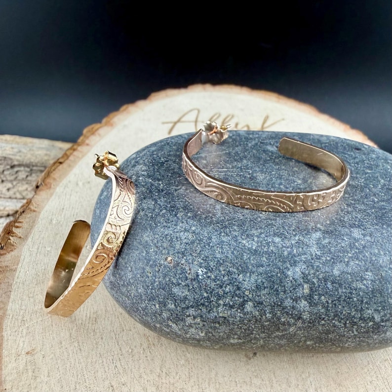 May include: A pair of gold-colored hoop earrings with an engraved floral design. The earrings are displayed on a gray stone, with a wooden background. The earrings have a post and are open at the bottom.