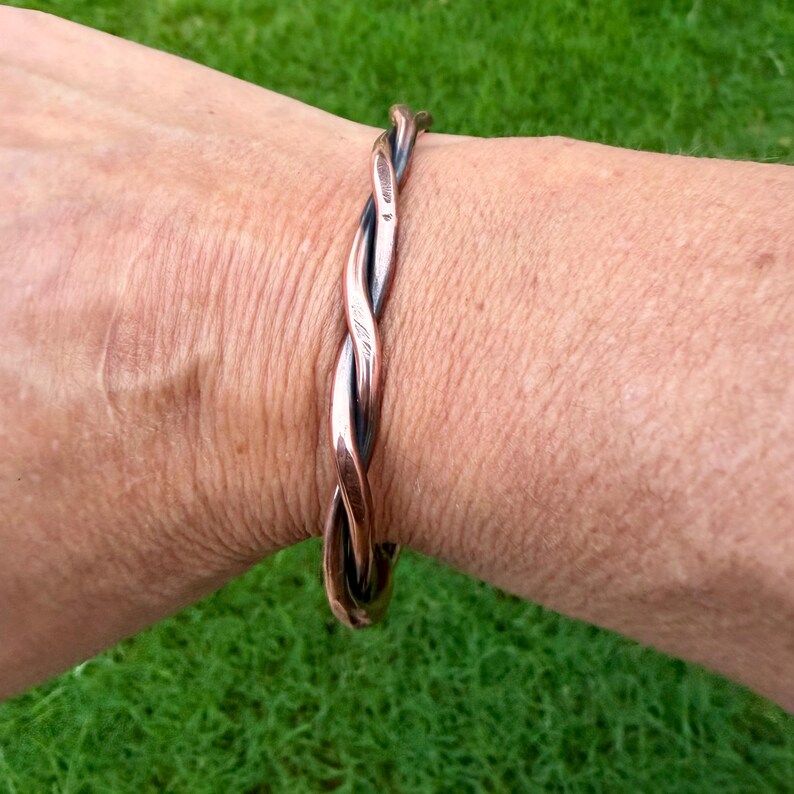 May include: A copper-colored, twisted metal bracelet. The bracelet has a braided design with a polished finish. The jewelry is worn on a wrist, with a green grassy background.