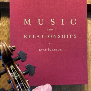 Könnte beinhalten: Ein rotes Hardcover-Buch mit dem Titel "MUSIC AND RELATIONSHIPS" in goldenen Buchstaben und dem Namen des Autors "EVAN JOHNSON" in kleineren goldenen Buchstaben.
