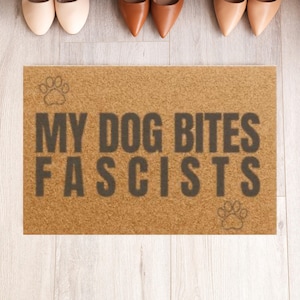 May include: A rectangular doormat with the text "MY DOG BITES FASCISTS" in dark gray letters. The mat is brown with a paw print at the top left and bottom right. Several pairs of shoes are visible above the mat.