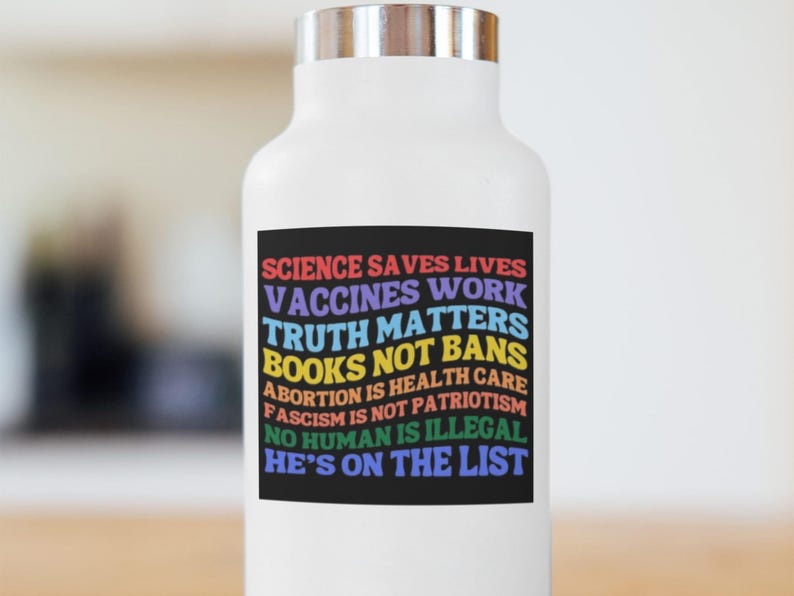 Pode incluir: Garrafa de &aacute;gua branca com tampa prateada. Um ret&acirc;ngulo preto na garrafa exibe texto colorido: "SCIENCE SAVES LIVES, VACCINES WORK, TRUTH MATTERS, BOOKS NOT BANS, ABORTION IS HEALTH CARE, FASCISM IS NOT PATRIOTISM, NO HUMAN IS ILLEGAL, HE'S ON THE LIST."