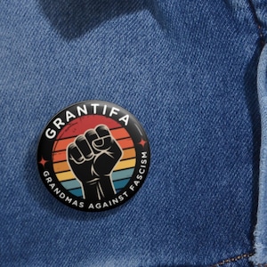 May include: A circular badge with the words "Grandmas Against Fascism" around a black fist graphic. The background has a retro sunset design with red, orange, yellow, and blue stripes. The badge is on a blue denim background.