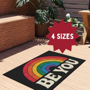 Puede incluir: Una alfombra rectangular negra con un diseño de arcoíris y las palabras "BE YOU" en blanco. La alfombra está sobre una terraza de madera, con un gráfico de estrella roja que dice "4 TAMAÑOS".