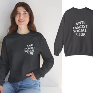 K&ouml;nnte beinhalten: Dunkelgraues Sweatshirt mit dem wei&szlig;en Text "ANTI FASCIST SOCIAL CLUB" auf der Vorder- und R&uuml;ckseite. Das lang&auml;rmlige Oberteil hat einen Rundhalsausschnitt. Das Model tr&auml;gt eine blaue Jeans.