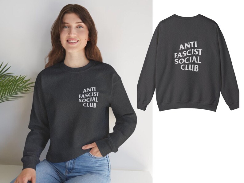K&ouml;nnte beinhalten: Dunkelgraues Sweatshirt mit dem wei&szlig;en Text "ANTI FASCIST SOCIAL CLUB" auf der Vorder- und R&uuml;ckseite. Das lang&auml;rmlige Oberteil hat einen Rundhalsausschnitt. Das Model tr&auml;gt eine blaue Jeans.