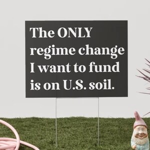 Op de afbeelding: Een zwart rechthoekig tuinbord met de tekst "The ONLY regime change I want to fund is on U.S. soil." wordt weergegeven op een groen gazon. Een roze tuinslang en een tuinkabouter staan op de voorgrond.