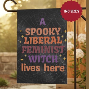 Può includere: Una bandiera da giardino nera con un disegno a ragnatela e la scritta "A Spooky Liberal Feminist Witch lives here" in arancione, viola e bianco. La bandiera è esposta all'aperto.