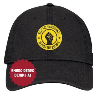 May include: Black denim hat with a yellow and black embroidered design. The design includes the text "KEEP THE IMMIGRANTS" and "DEPORT THE RACISTS" around a clenched fist symbol. The hat also has a red sticker that says "EMBROIDERED DENIM HAT".