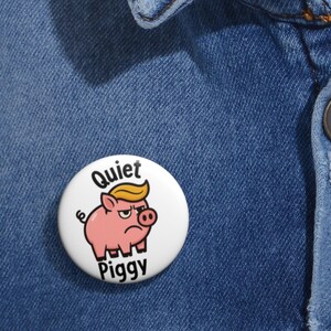 May include: A white button pin with a cartoon pig wearing a blonde wig and the words "Quiet Piggy". The pin is set against a blue denim background. The pig has an angry expression.