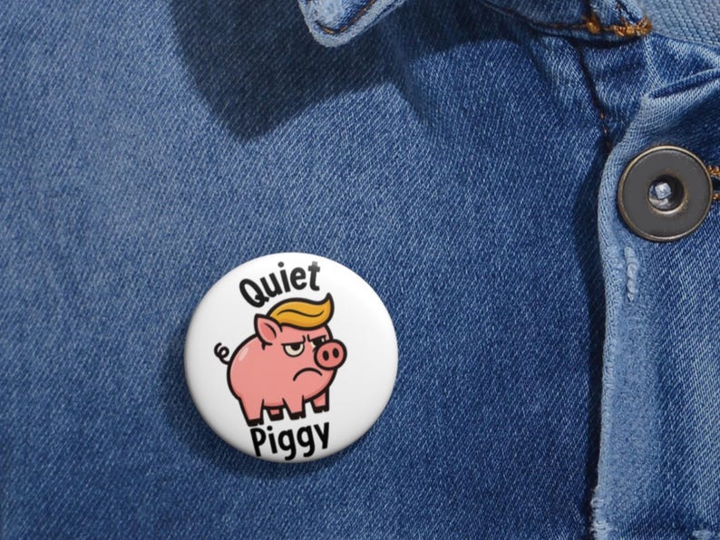 May include: A white button pin with a cartoon pig wearing a blonde wig and the words "Quiet Piggy". The pin is set against a blue denim background. The pig has an angry expression.