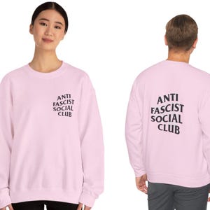 K&ouml;nnte beinhalten: Hellrosa Sweatshirt mit dem Text "ANTI FASCIST SOCIAL CLUB" in Schwarz auf der Vorder- und R&uuml;ckseite. Das Sweatshirt hat einen Rundhalsausschnitt.