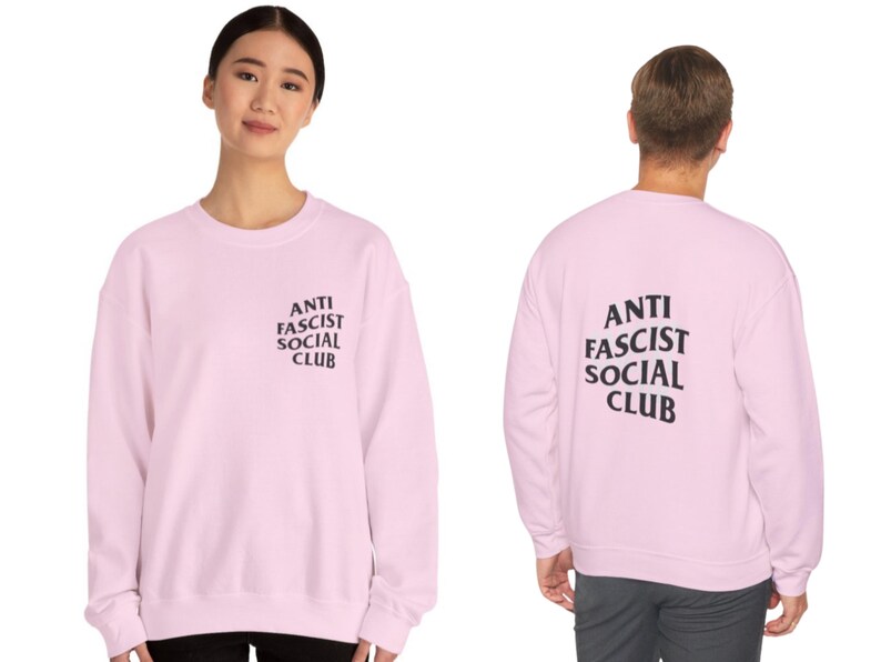 K&ouml;nnte beinhalten: Hellrosa Sweatshirt mit dem Text "ANTI FASCIST SOCIAL CLUB" in Schwarz auf der Vorder- und R&uuml;ckseite. Das Sweatshirt hat einen Rundhalsausschnitt.